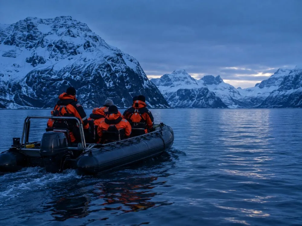 Passengers in thermal suits gripping the handles of an open RIB as it crosses a dark fjord.